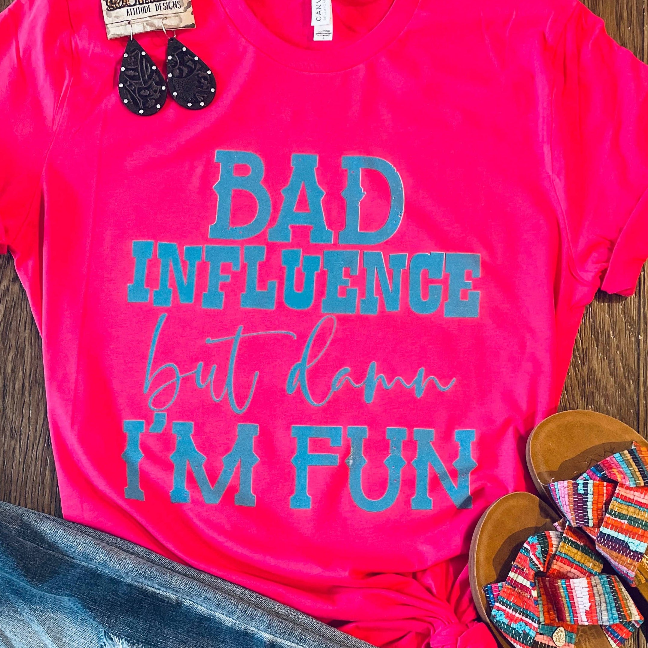 Pink t-shirt with 'Bad Influence but damn I'm fun' text, denim jeans, and sandals on a wooden surface.