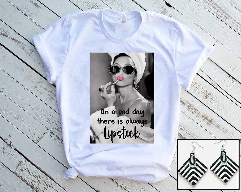 White t-shirt with graphic design and text on a wooden surface, accompanied by black and white earrings.