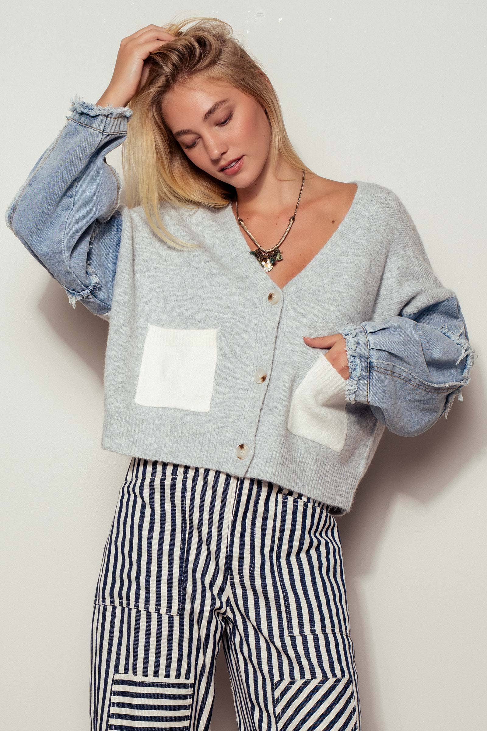 Woman wearing a gray cardigan with a white pocket, blue jeans, and striped pants against a plain background.