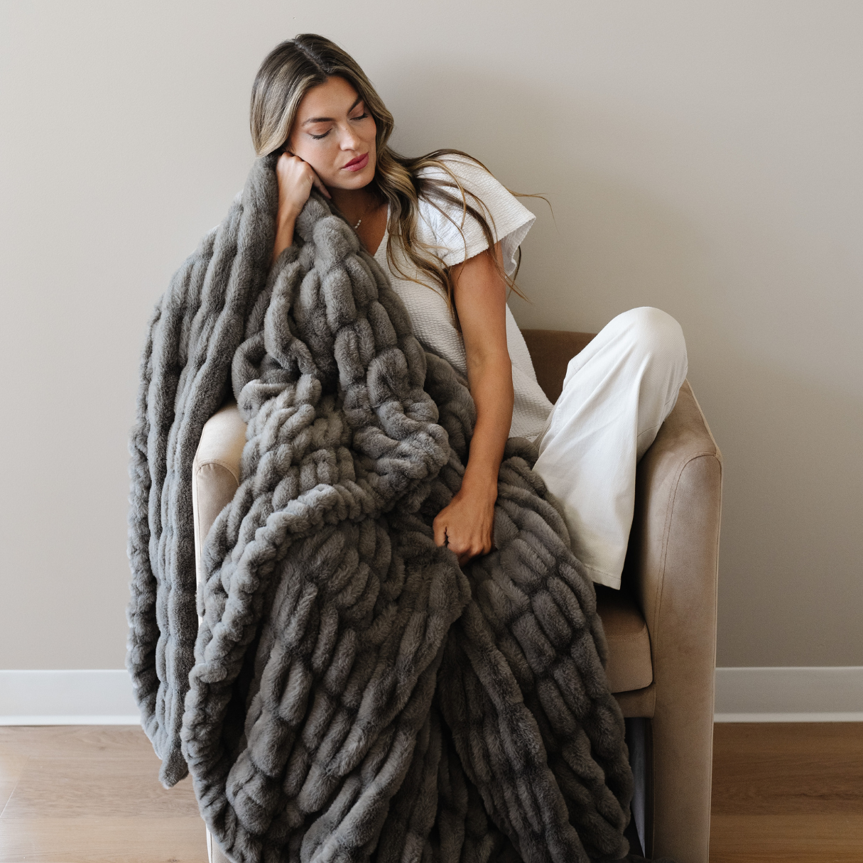 Woman sitting on a couch wrapped in a large gray blanket in a cozy room.