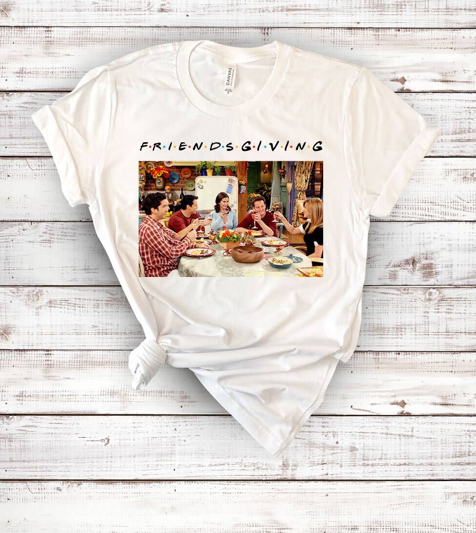 White t-shirt with 'Friendsgiving' graphic featuring a group of people around a table on a wooden surface.