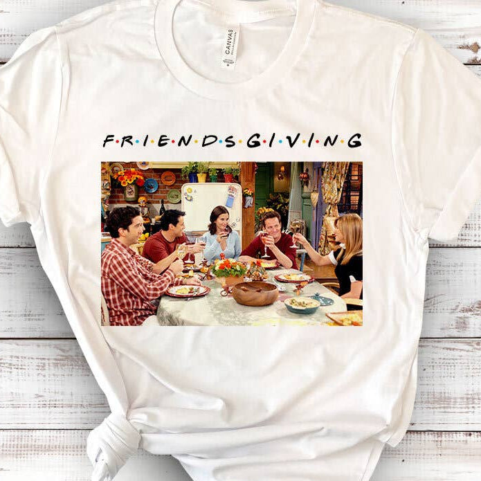 White t-shirt with 'Friendsgiving' graphic featuring a group of people around a table on a wooden surface.