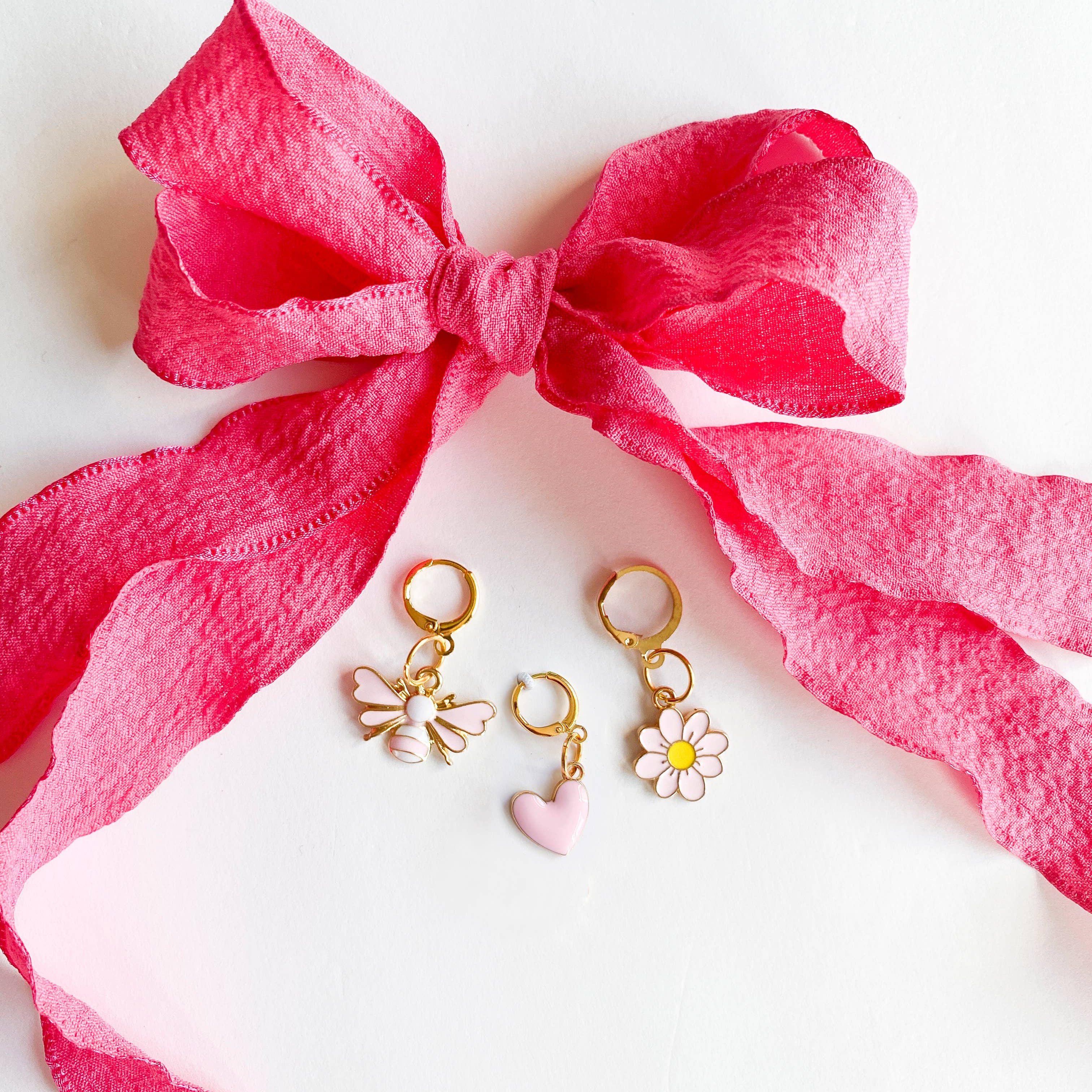 Two gold earrings with butterfly and flower charms on a white surface with a pink bow.