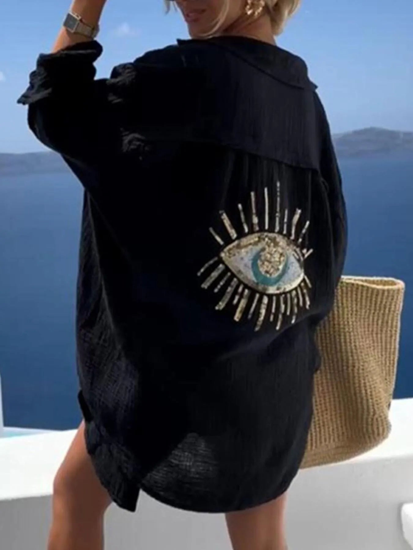 Person wearing a black jacket with an embroidered eye design, standing against a scenic background.