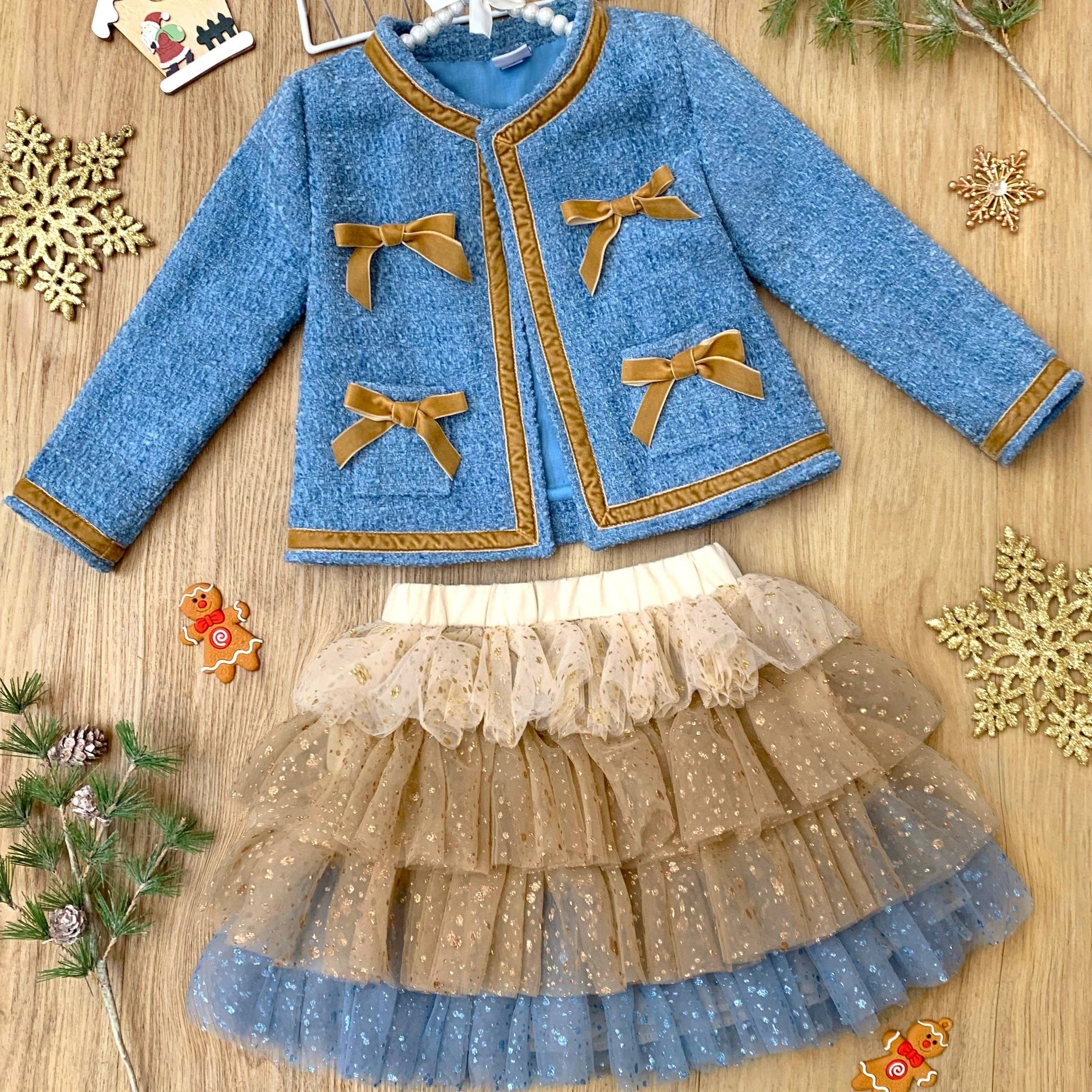 Children's blue jacket with gold bows and beige skirt on a wooden surface with decorative items.