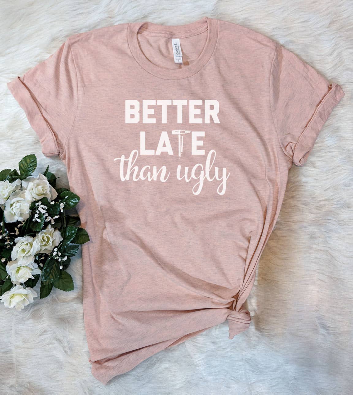 Pink t-shirt with 'Better Late than Ugly' text on a white surface with flowers.