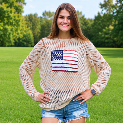 Woman wearing a beige mesh top with an American flag design in a grassy field.
