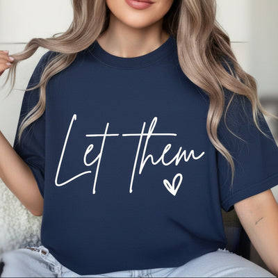 Person wearing a black t-shirt with 'Let Them' text and heart symbol, sitting on a couch.