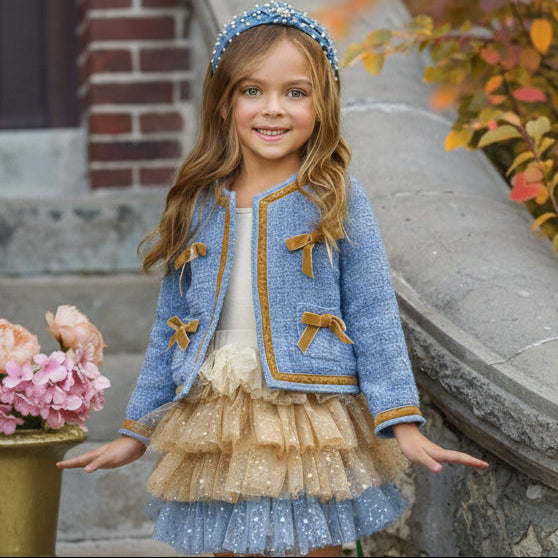 Young girl wearing a blue jacket with gold accents and a beige skirt, standing outdoors.