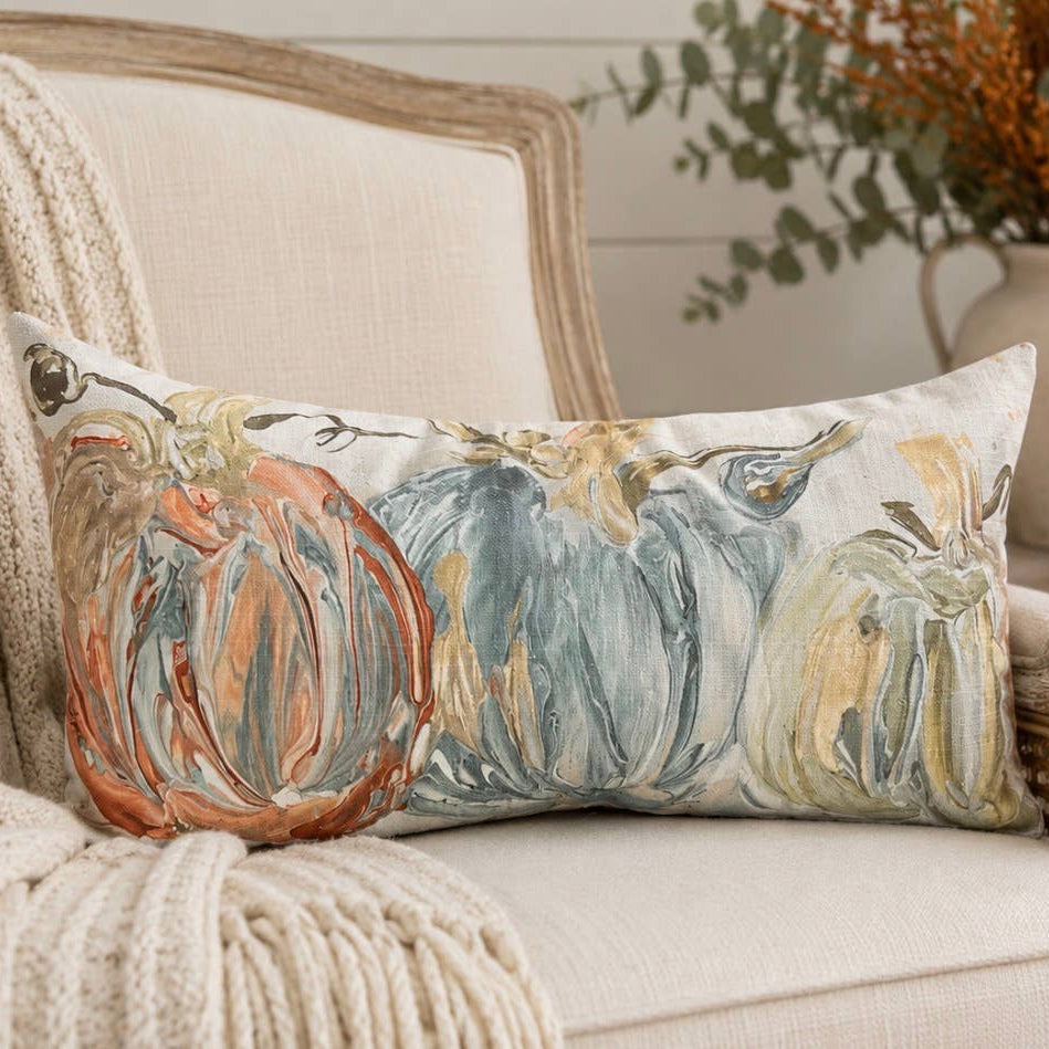 A rectangular decorative throw pillow with a painted pumpkin design in muted orange, blue-gray, and beige tones, resting on a cream upholstered armchair in a styled indoor setting with a knit blanket, candle, and vase in the background.