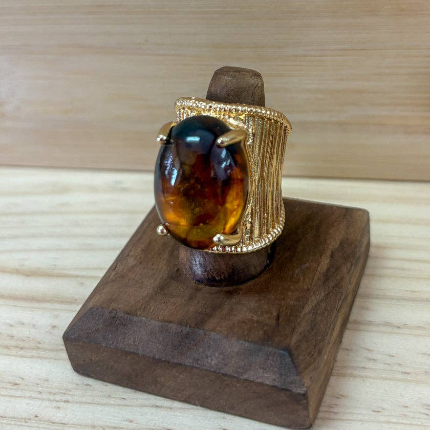 A gold ring with a large oval amber cabochon stone, displayed on a dark wooden stand over a light wood surface.