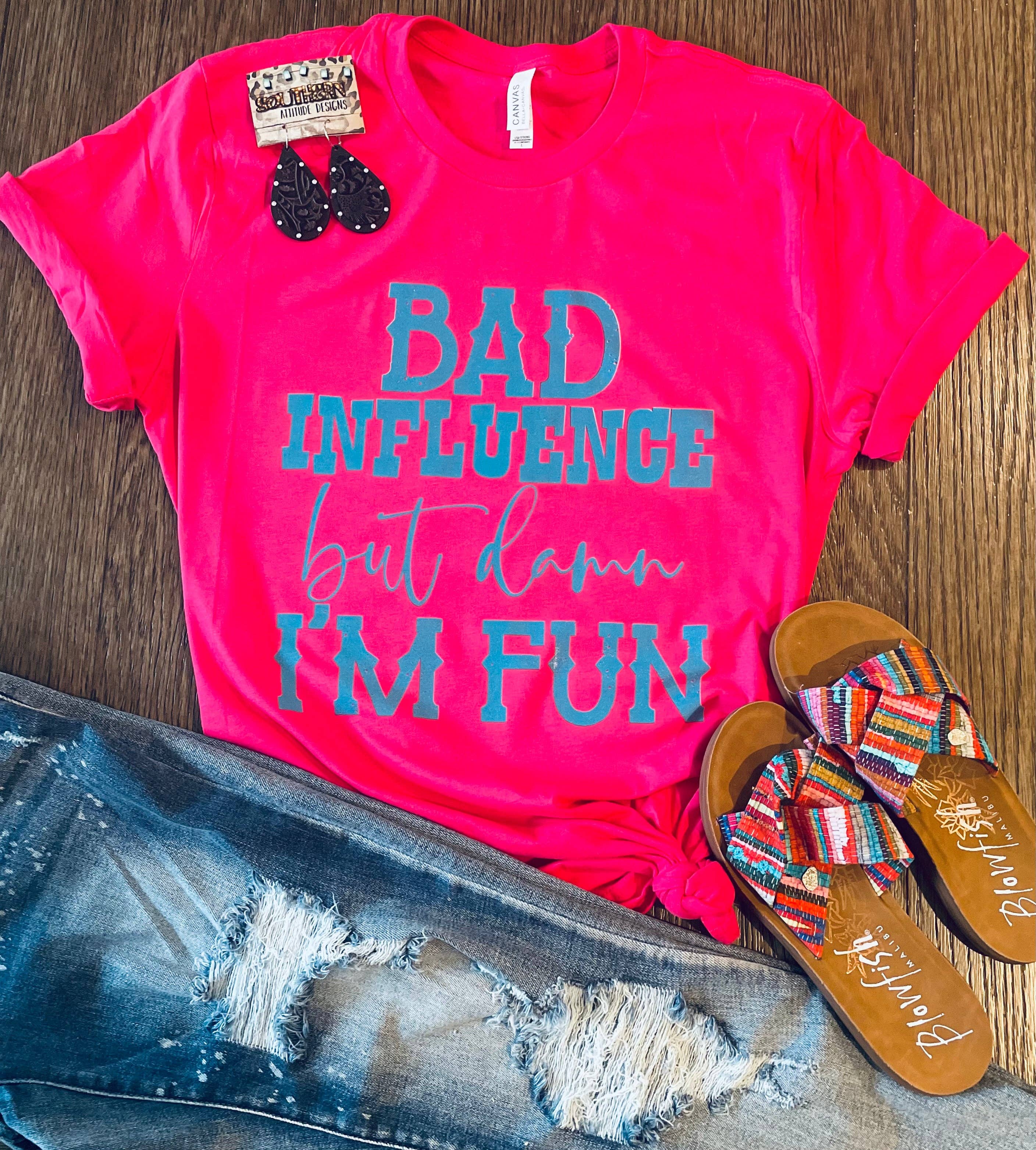 Pink t-shirt with 'Bad Influence but damn I'm fun' text, denim jeans, and sandals on a wooden surface.