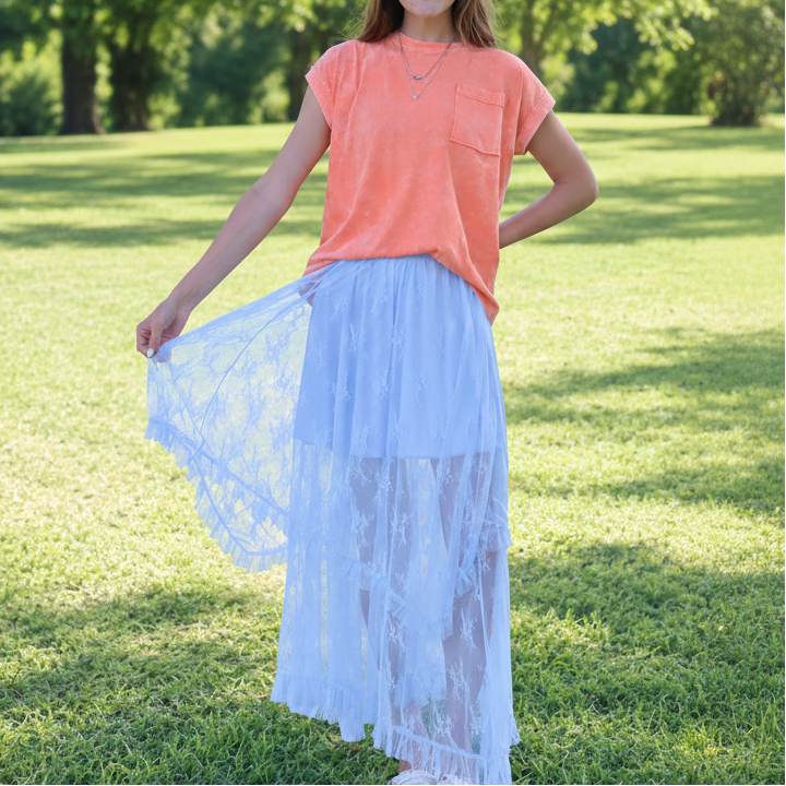 Woman wearing a coral top and light blue lace skirt standing in a grassy field.