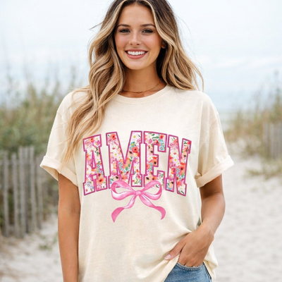 Woman wearing a cream t-shirt with floral letters and a pink bow on a beach.