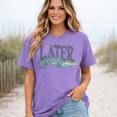 Woman wearing a purple t-shirt with an alligator graphic and 'LATER' text on a beach.