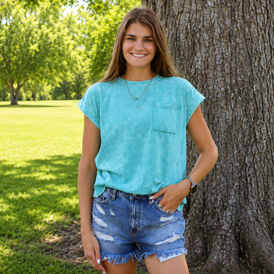 Woman in a turquoise shirt and denim shorts standing next to a large tree in a park.