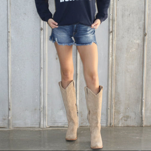 Person wearing a navy sweatshirt with 'HE'S HUNTING' text, denim shorts, and tan boots against a gray wall.