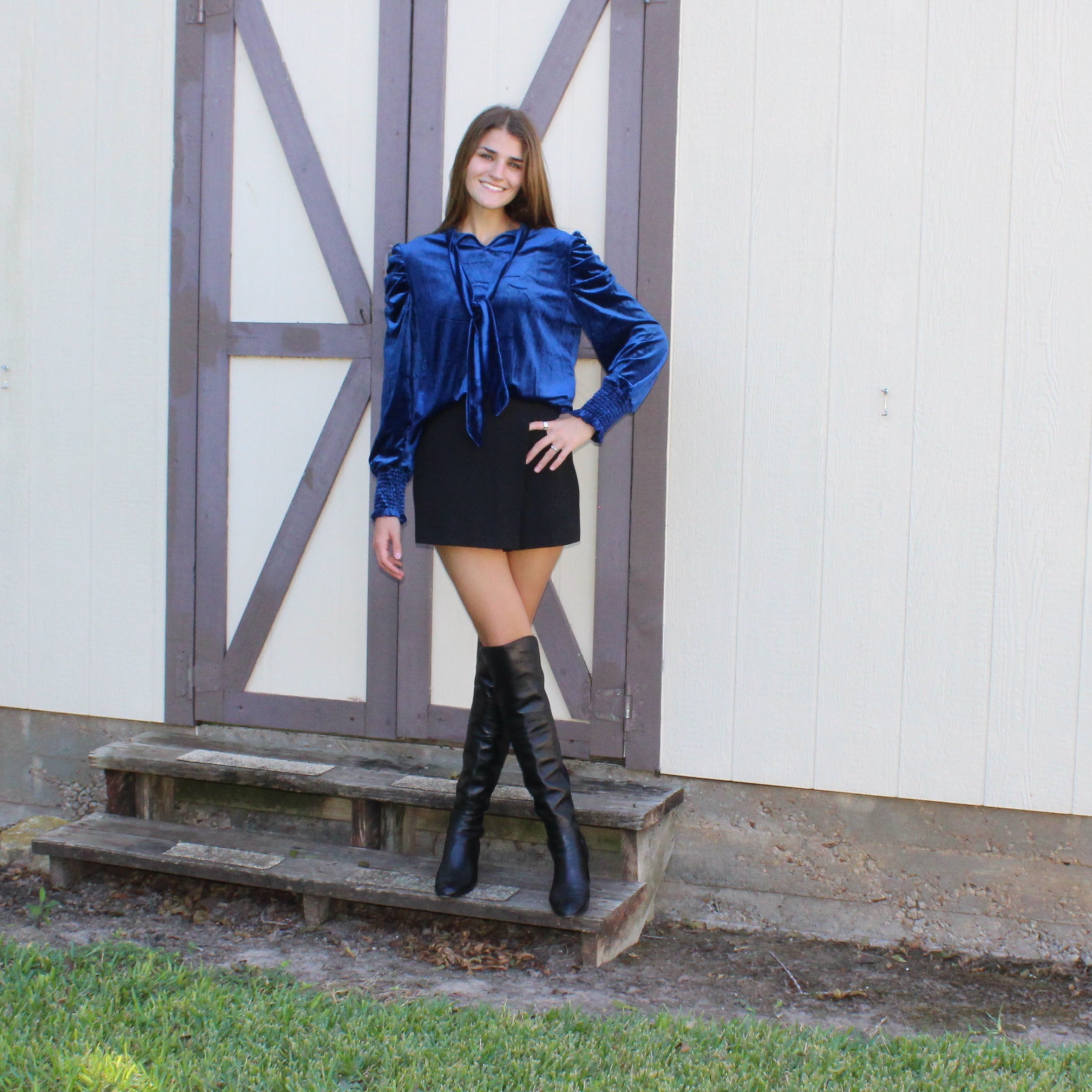Woman in a blue velvet jacket and black skirt standing in front of a small building with a wooden door.