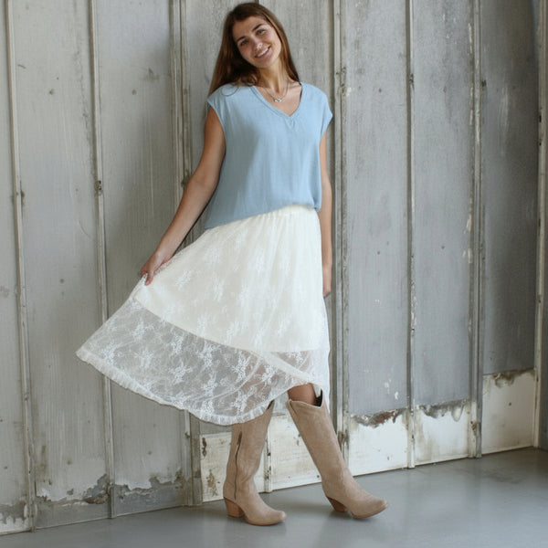 Woman in a light blue top and white lace skirt standing in a rustic room with wooden walls.