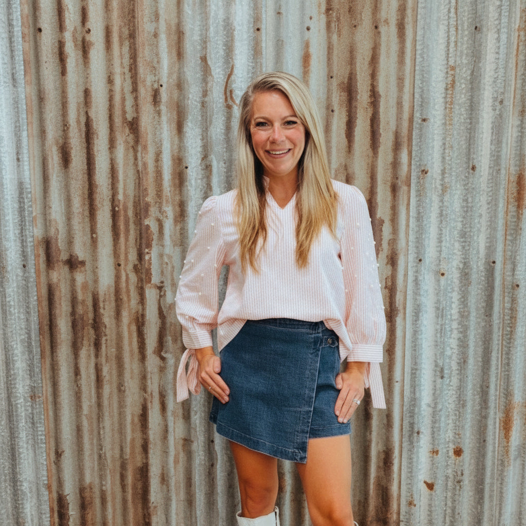 Woman standing in front of a decorative wall with text and a coat, wearing a pink blouse, denim skirt, and white boots.