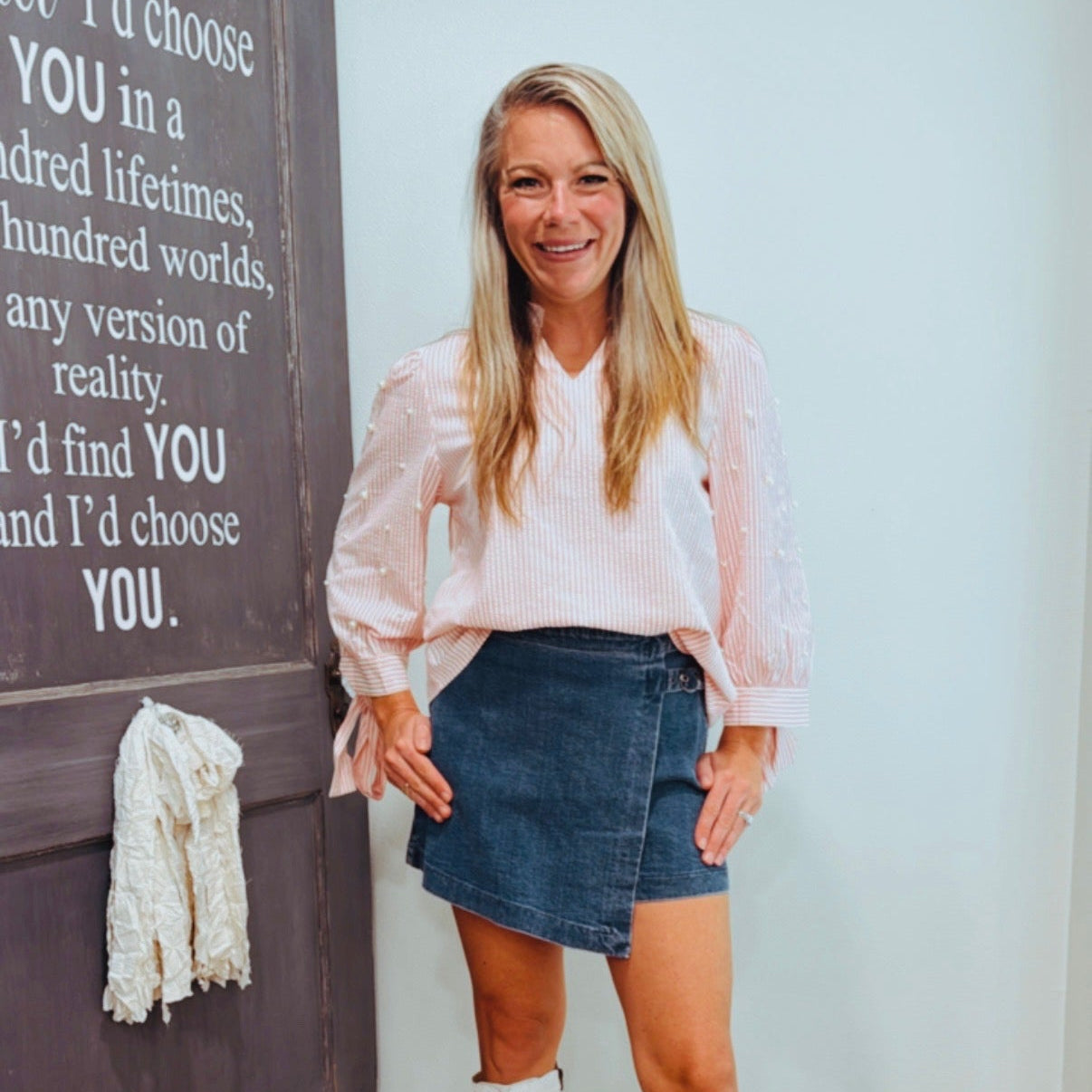 Woman standing in front of a decorative wall with text and a coat, wearing a pink blouse, denim skirt, and white boots.
