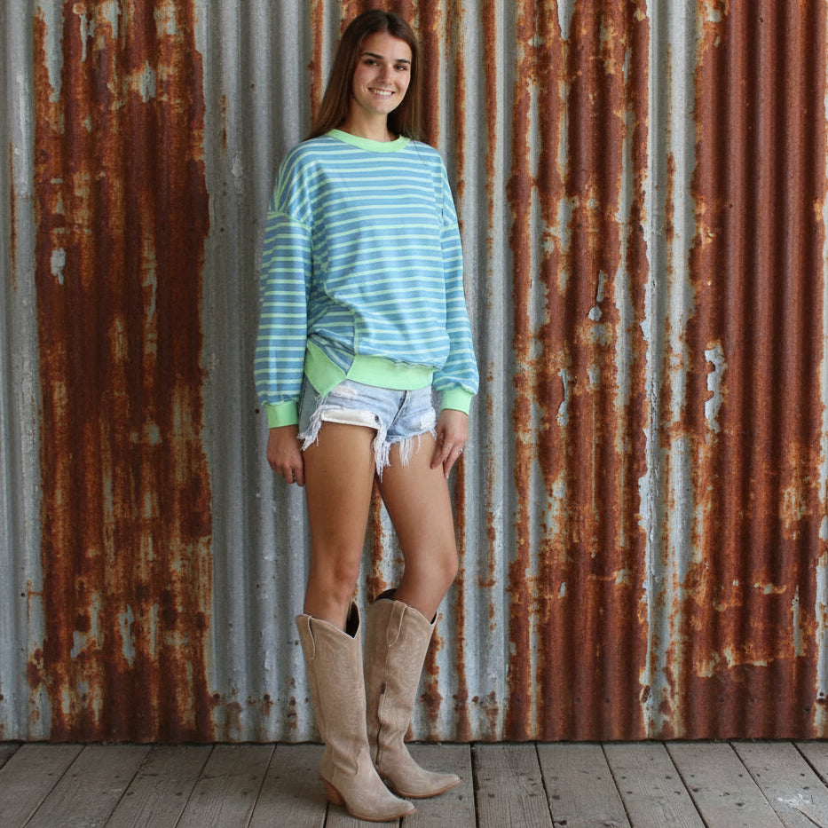 Woman wearing a striped sweater and shorts with boots standing against a wooden wall.