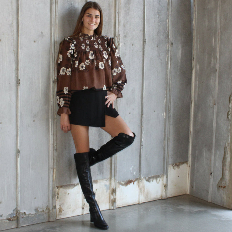 Woman wearing a floral blouse and black skirt with knee-high boots against a rustic wooden wall.