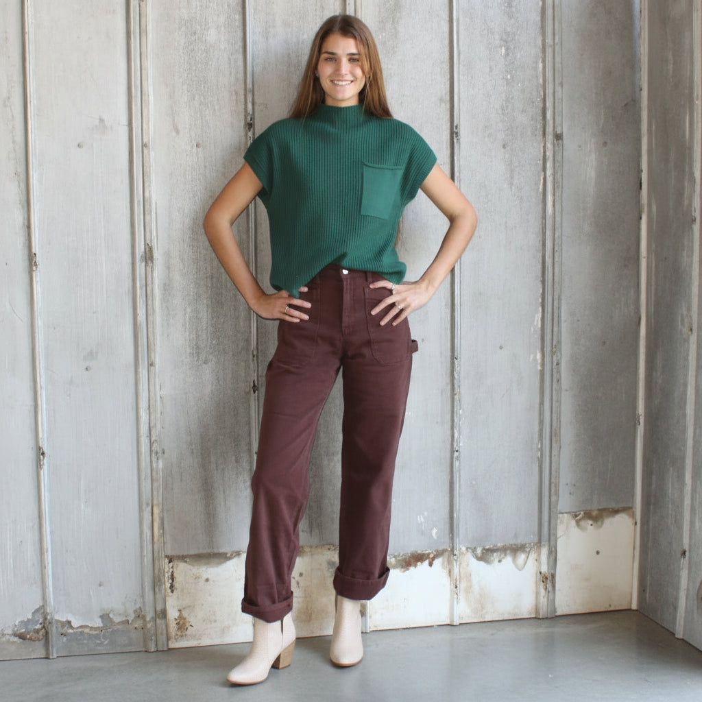 Woman wearing a green top and maroon pants standing against a wooden wall.
