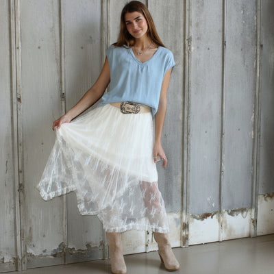 Woman wearing a light blue top and white lace skirt standing in a rustic wooden room.