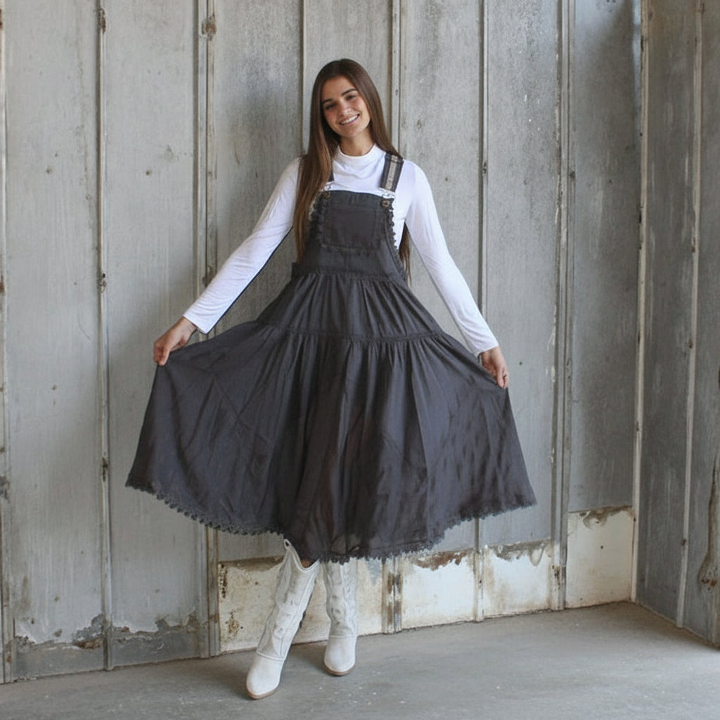 Woman wearing a black pinafore dress with a white long-sleeve shirt against a wooden wall.