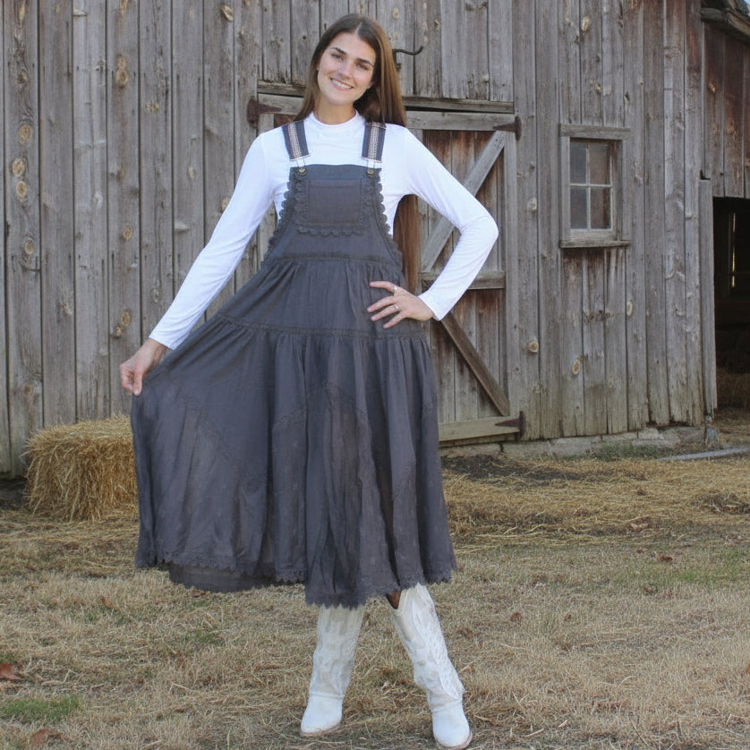 Woman wearing a long dress with suspenders in front of a wooden barn.