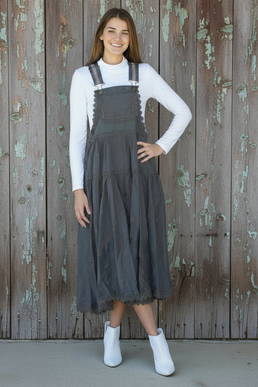 Woman wearing a dark denim pinafore dress over a white long-sleeve shirt against a wooden background.