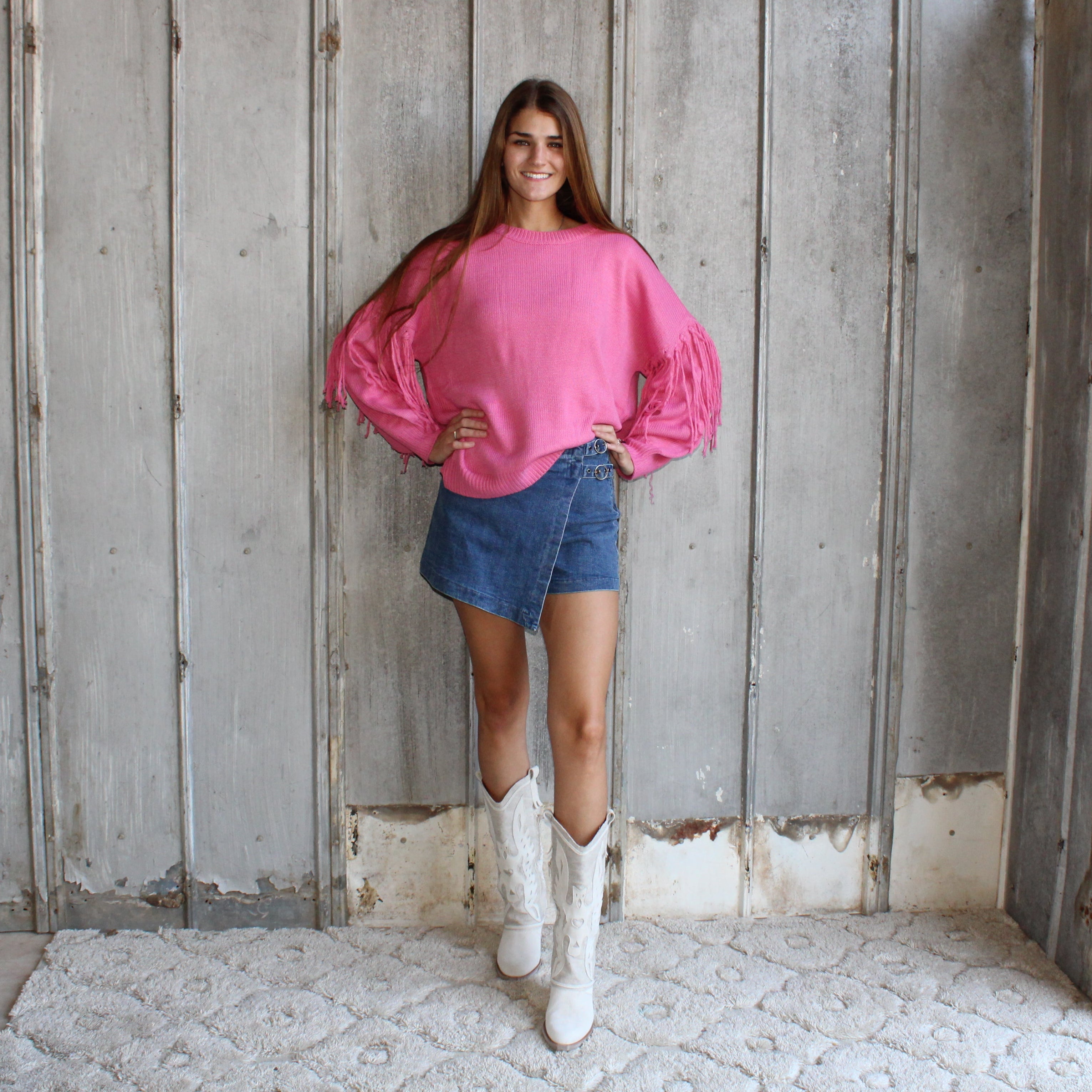 Woman wearing a pink sweater and denim shorts standing in a rustic room with wooden walls.