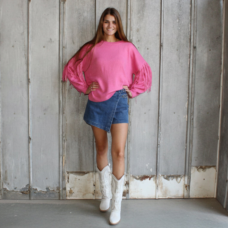 Woman wearing a pink sweater and denim shorts standing in a rustic room with wooden walls.