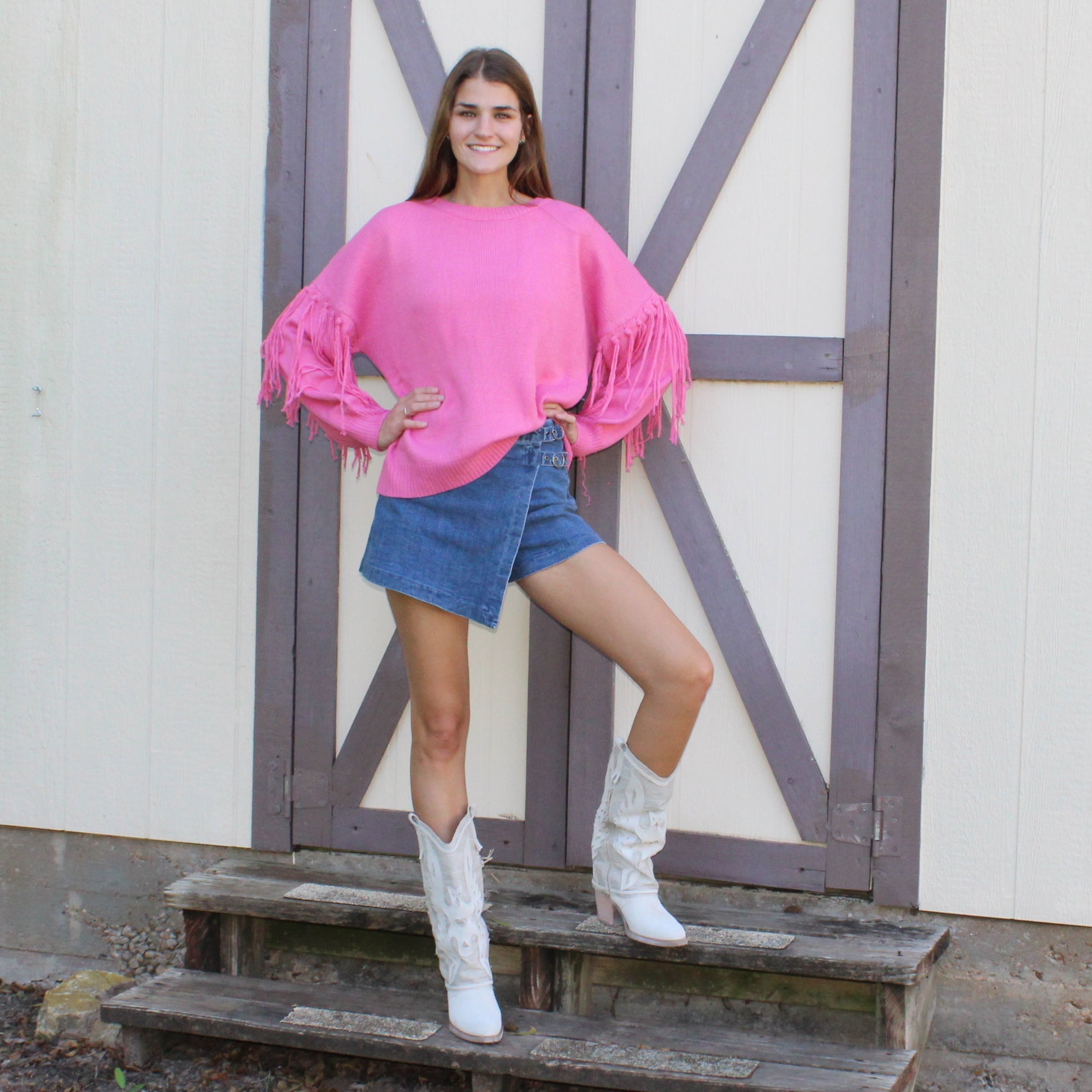 Woman in pink sweater and denim shorts standing on a wooden staircase in front of a white building with gray doors.