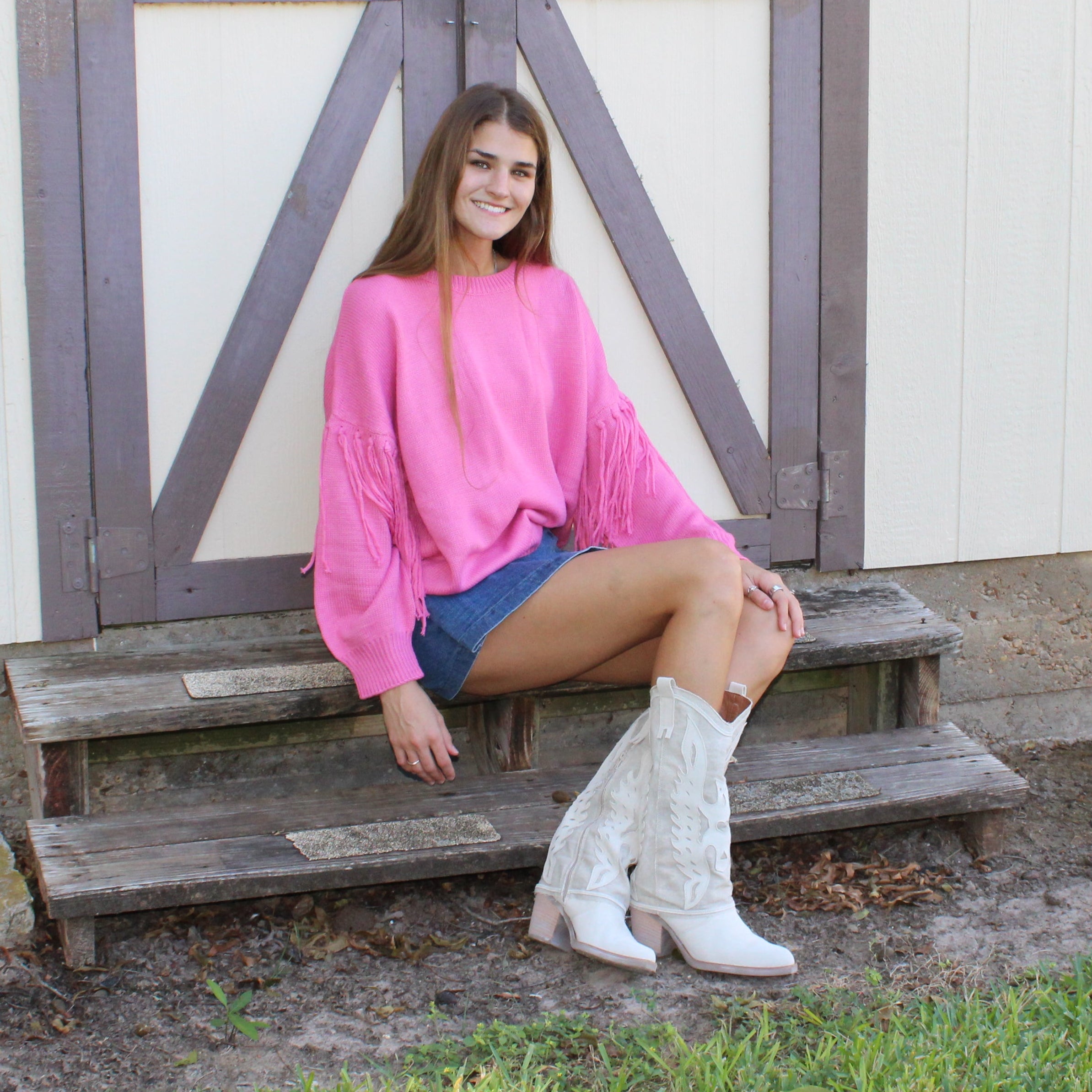 Woman in pink sweater and white boots sitting on a wooden bench in front of a white building with gray doors.