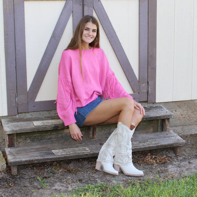 Woman in pink sweater and white boots sitting on a wooden bench in front of a white building with gray doors.