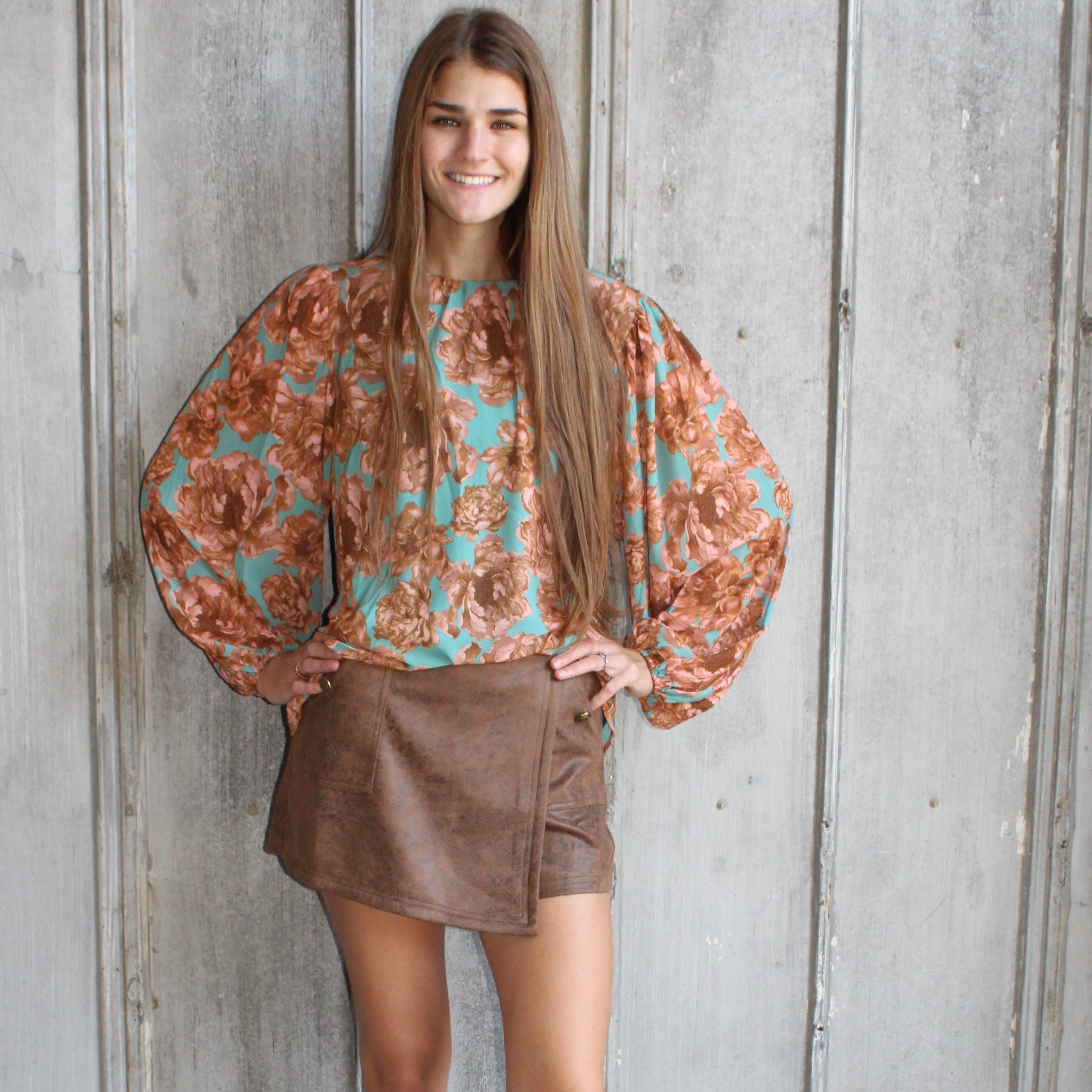 Woman wearing a floral top and brown skirt standing against a textured wall.