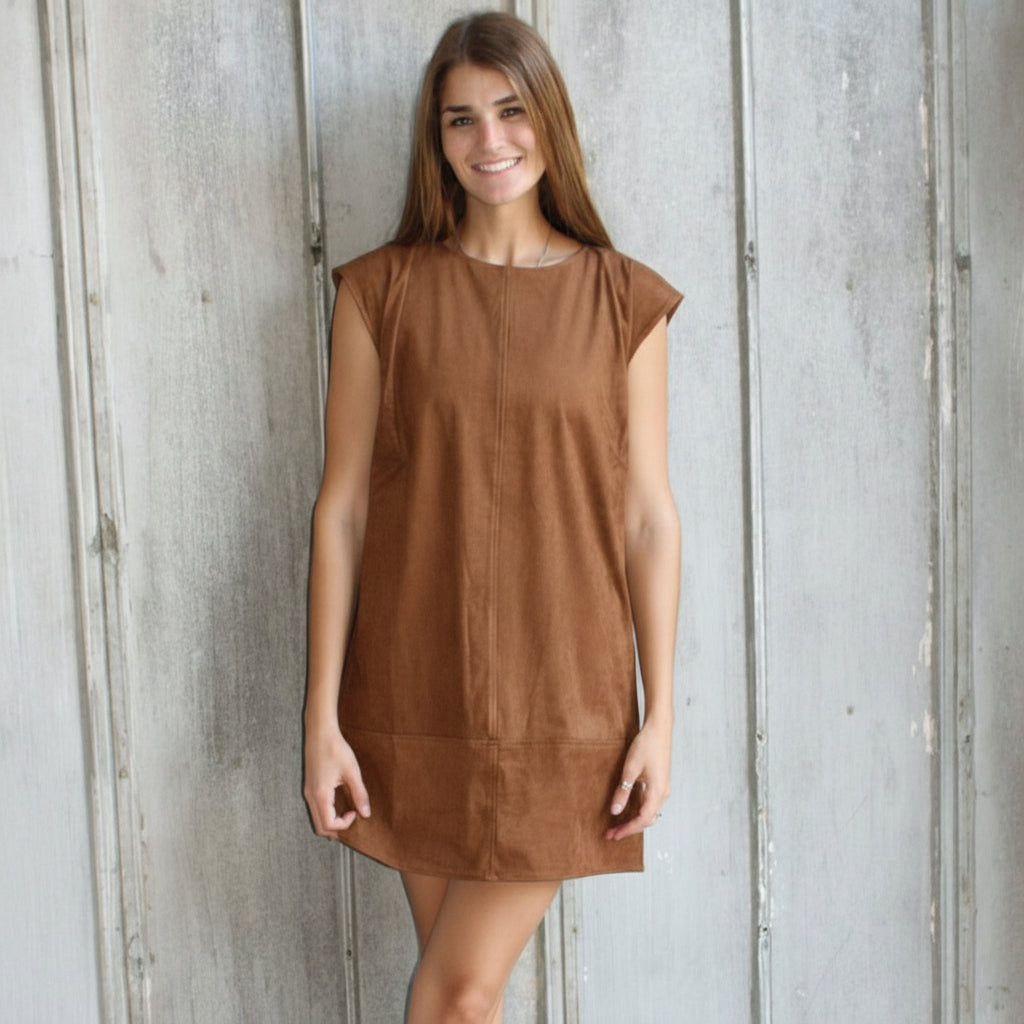 Woman in a brown dress and white boots standing against a textured wall.