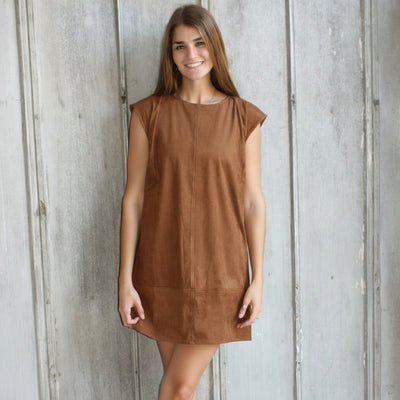 Woman in a brown dress and white boots standing against a textured wall.