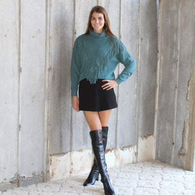 Woman in teal sweater and black skirt standing on a wooden staircase.
