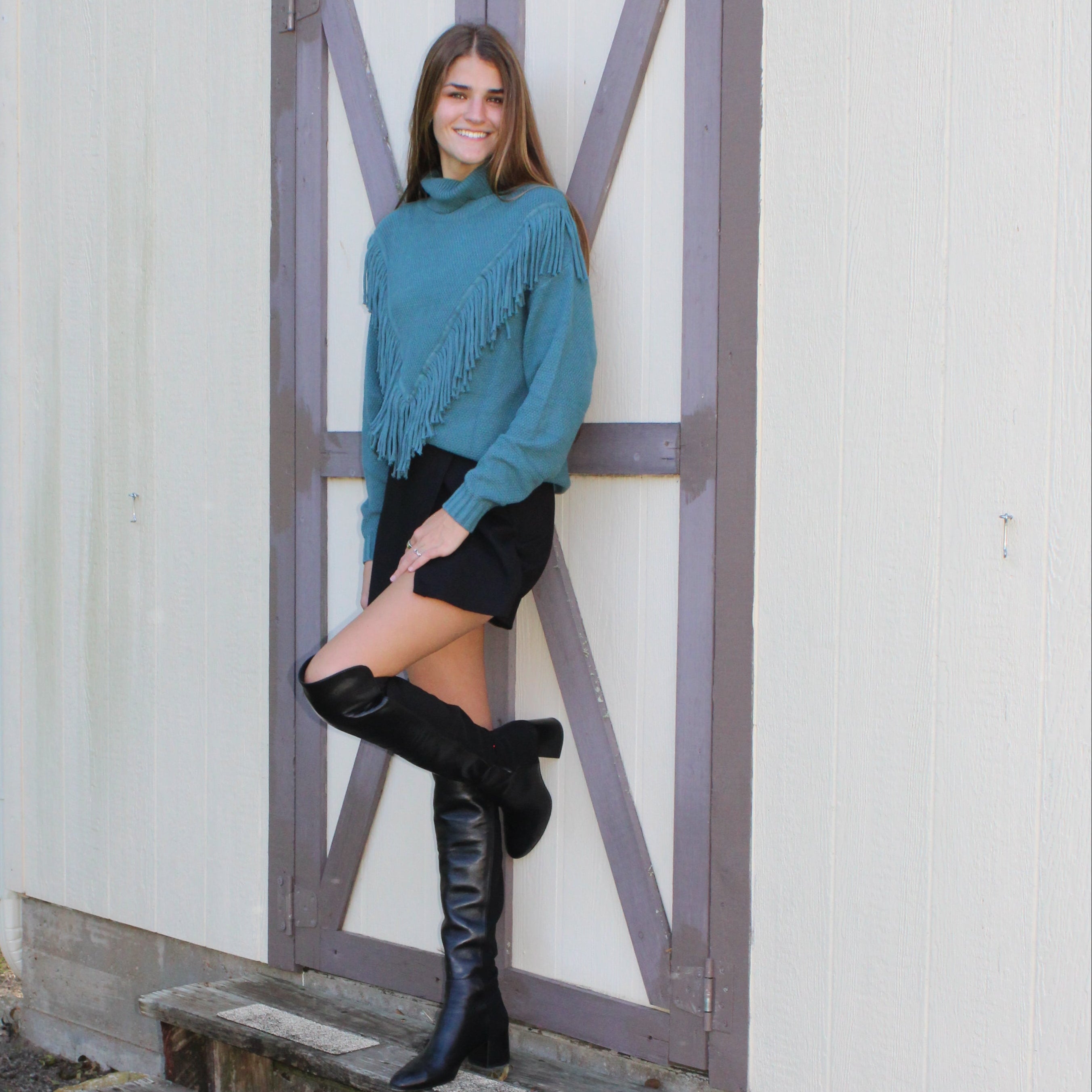 Person standing in front of a small wooden structure with a white wall and grassy area in the background.