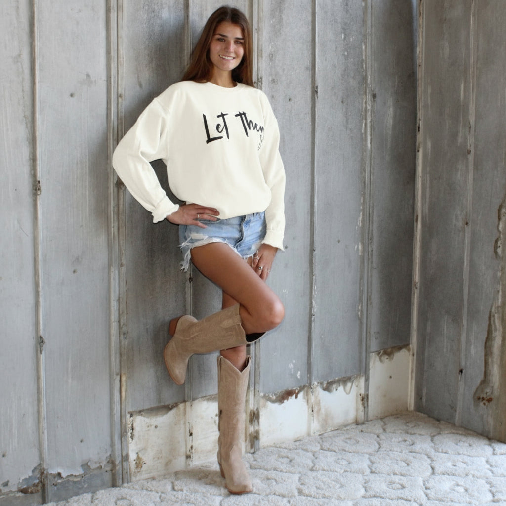 Woman in a white sweatshirt and denim shorts standing against a wooden wall.