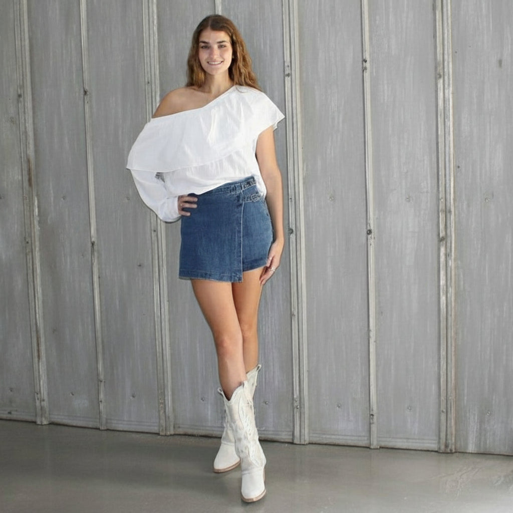 Woman in a white top and denim skirt standing in front of a wooden wall.