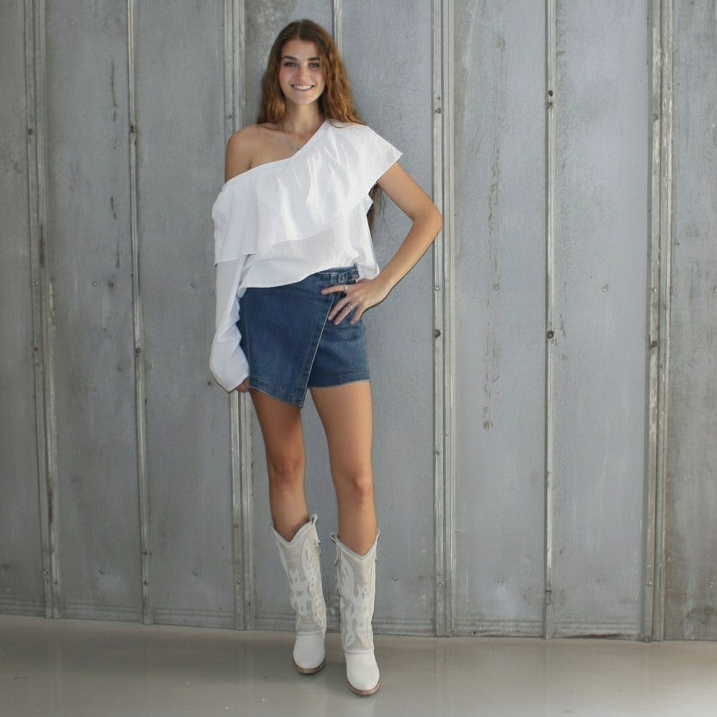 Woman in white off-shoulder top and denim shorts standing against a gray wooden wall.