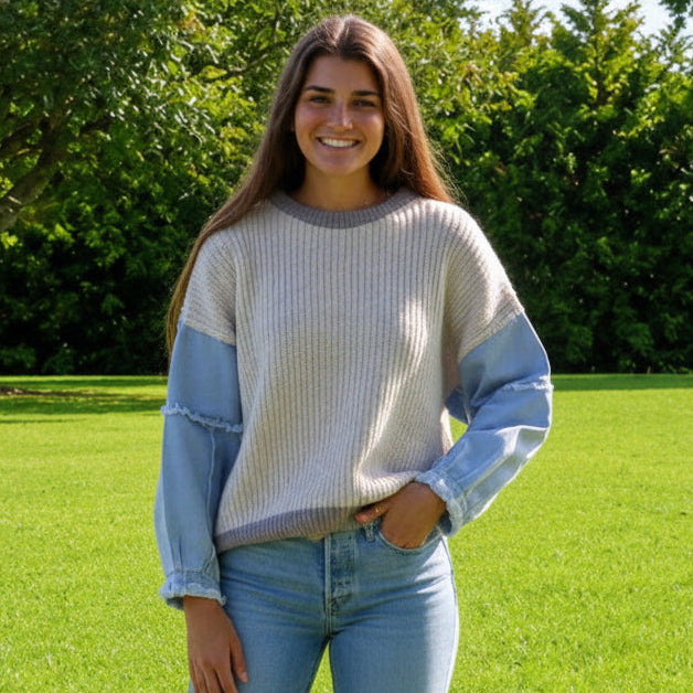 A young woman stands barefoot on a bright green lawn in a park, wearing a color-blocked knit sweater with a cream body and light blue sleeves, paired with denim shorts.