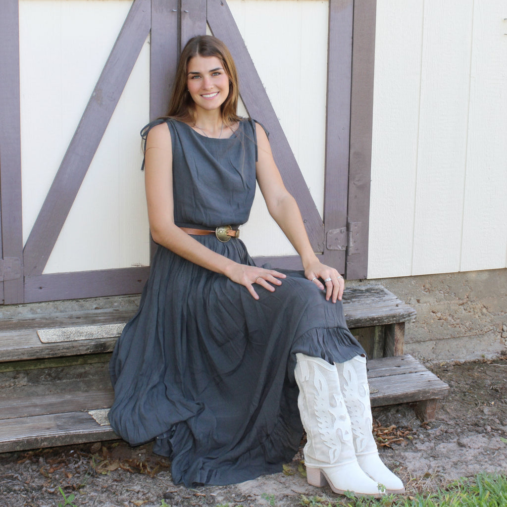 Woman in a long dark dress and white boots sitting on a wooden bench in front of a white building with a gray door.