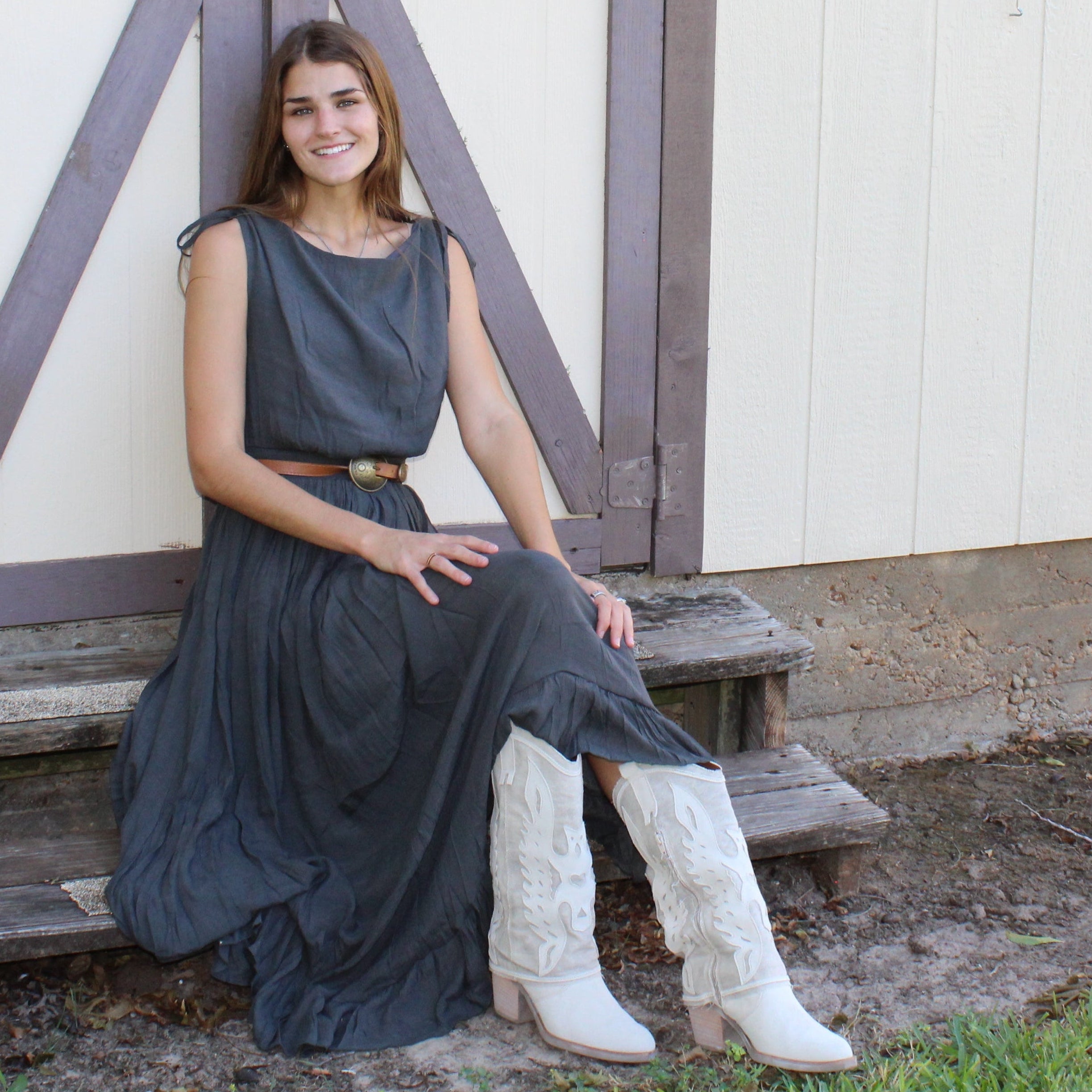 Woman in a long dark dress sitting on a wooden bench against a white building with wooden doors.