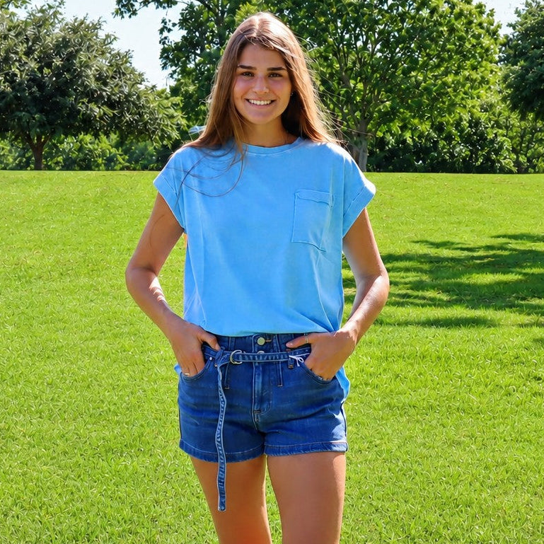 A young woman stands on a green grassy lawn wearing a light blue short-sleeve t-shirt and denim shorts, with trees in the background.