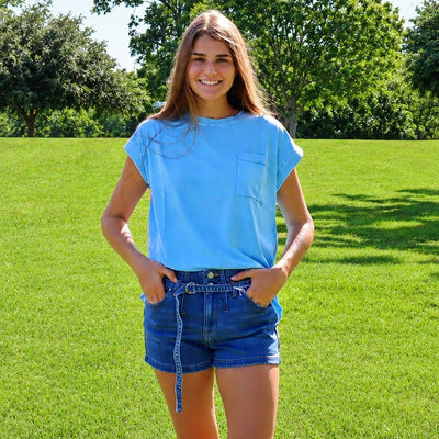 A young woman stands on a green grassy lawn wearing a light blue short-sleeve t-shirt and denim shorts, with trees in the background.